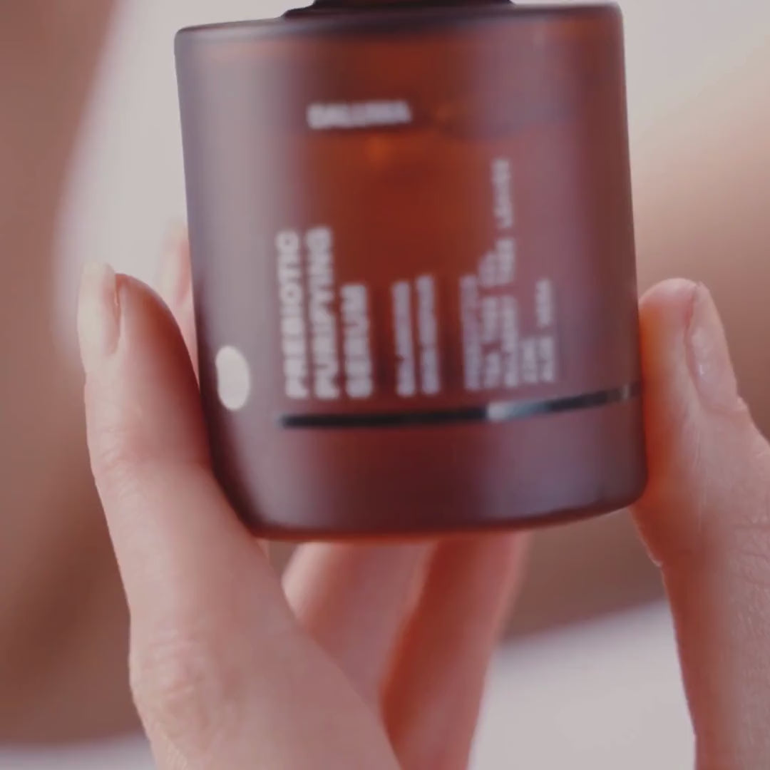 Eine Hand hält einen braunen, gefrosteten Tiegel mit der Aufschrift Prebiotic Purifying Cream, wobei der Text teilweise sichtbar ist; der Hintergrund ist leicht unscharf.
