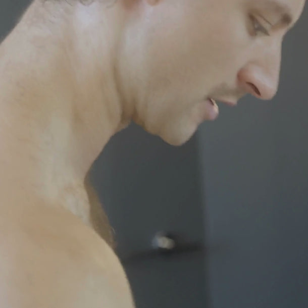 Eine Nahaufnahme des Seitenprofils eines hemdlosen Mannes mit kurzen Haaren, der mit einem neutralen Gesichtsausdruck nach unten schaut und Dalumas GREEN COFFEE CLEANSER ENZYMPEELING vor einem unscharfen Hintergrund zeigt.