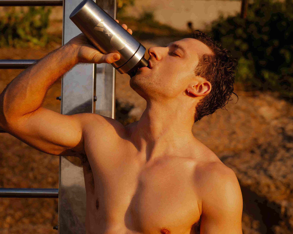 Ein Mann ohne Hemd und mit kurzen Haaren trinkt im Freien aus einer metallischen Wasserflasche, wobei das Sonnenlicht sein Gesicht und seinen Körper beleuchtet.