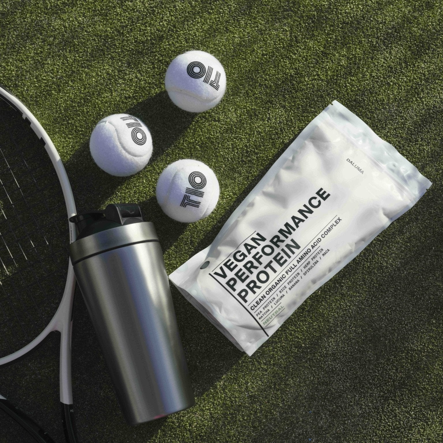 Eine Shakerflasche, eine Packung mit der Aufschrift VEGANES PERFORMANCE PROTEIN KAKAO DOUBLE von DALUMA, drei Tennisbälle und ein Teil eines Tennisschlägers liegen auf grünem Kunstrasen im Sonnenlicht.