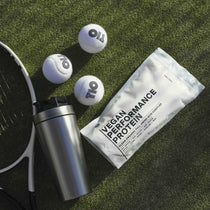Eine Shakerflasche, eine Packung mit der Aufschrift VEGANES PERFORMANCE PROTEIN KAKAO DOUBLE von DALUMA, drei Tennisbälle und ein Teil eines Tennisschlägers liegen auf grünem Kunstrasen im Sonnenlicht.
