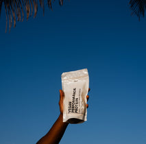 Eine Hand hält eine weiße DALUMA VEGANES PERFORMANCE PROTEIN NEUTRAL-Tüte vor einem klaren blauen Himmel hoch, in der linken oberen Ecke sind Palmenblätter zu sehen.