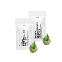 Zwei weiße DALUMA VEGANES PERFORMANCE PROTEIN NEUTRAL DOUBLE-Beutel stehen aufrecht, jeder mit einem Haufen grünem pflanzlichem Proteinpulver und zwei grünen Blättern davor - ideal für veganen Muskelaufbau - vor weißem Hintergrund.